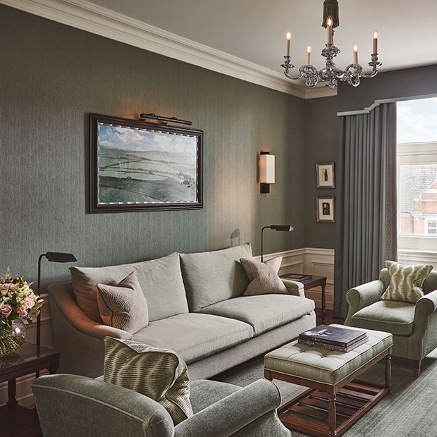 Coburg Suite: Living area of suite with sofa and cushions on the left, and a painting on the wall. A coffee table with books in front of the sofa, and a chair with a green cushion. The colours are shades of green.