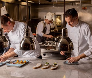 Chefs in white uniforms meticulously plating gourmet dishes under heat lamps in a professional kitchen, with two chefs in the foreground focusing on presentation and two more cooking in the background.