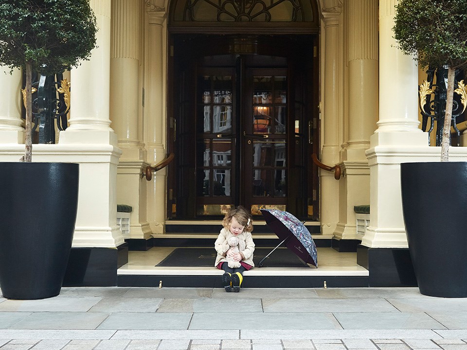 Photo of a playful little girl in front of the entrance, playing with her teddy bear.