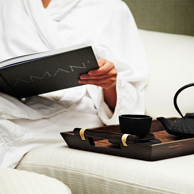 A girl in a bathrobe, holding an Aman Spa book and next to her is a teapot and a cup on a tray.