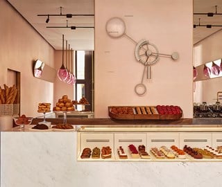 The patisserie counter with pastries, fresh baguettes, and croissants, framed by a soft pink interior and a large wall clock.