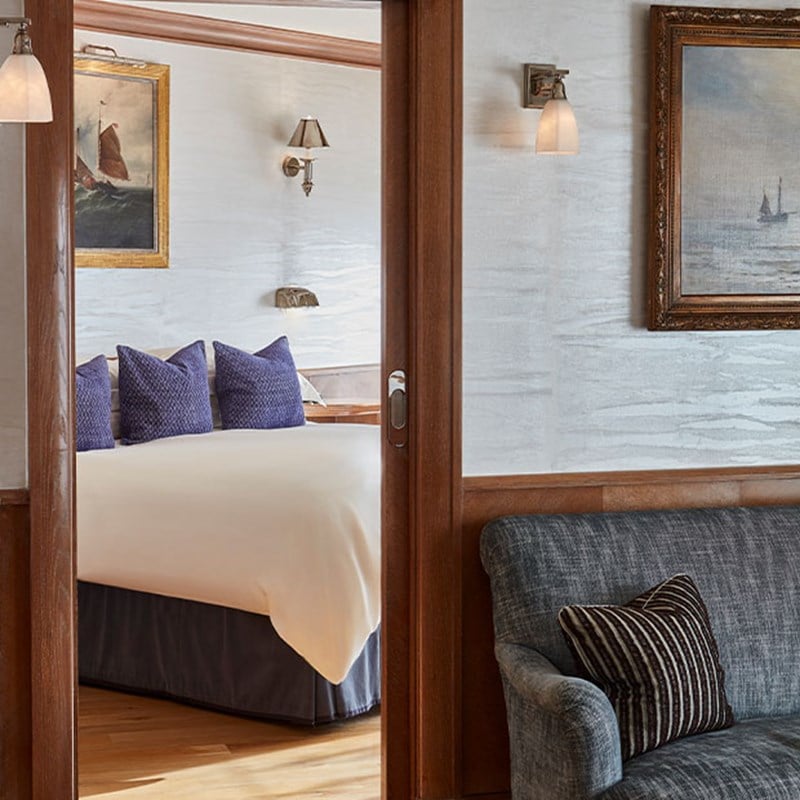 View through wooden sliding doors into a bedroom with a neatly made double bed, deep blue cushions, and maritime-themed paintings on the walls. A grey armchair and wall sconce appear in the foreground.