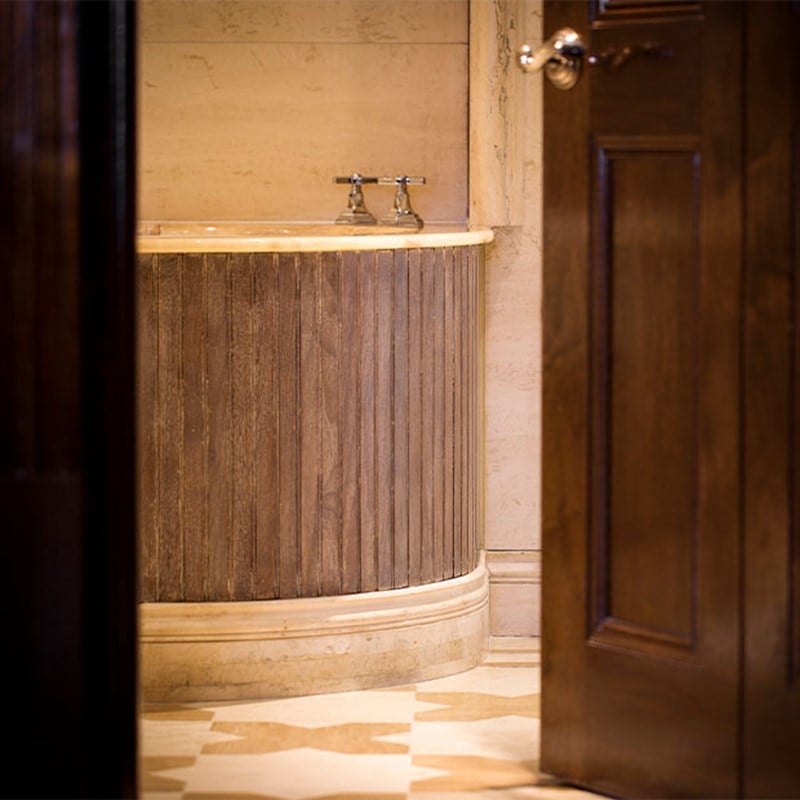 Close-up of a freestanding bathtub with ribbed wooden cladding and polished stone rim, set in a marble bathroom and partially visible through an open wooden door.