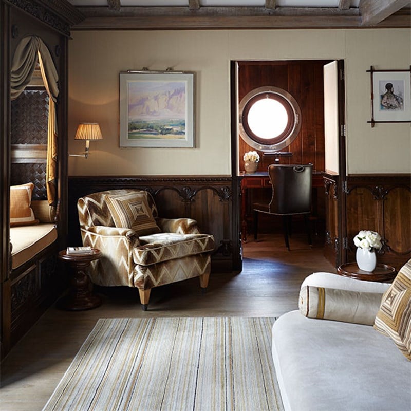 Living space with a patterned armchair, window seat, and writing nook framed by dark wood and a round porthole-style window. Soft lighting and artwork contribute to the refined, heritage feel.