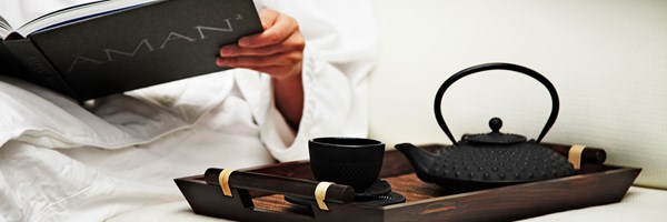 Woman holding Aman book in a bathrobe, whilst enjoying a tea on a wooden tray