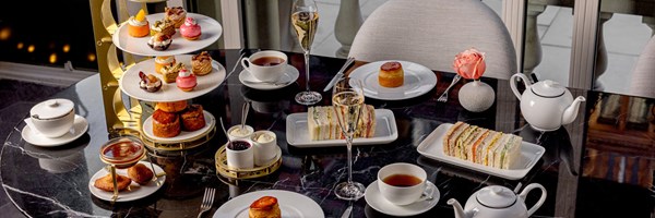 An elegant afternoon tea setting on a black marble table, adorned with fine china, a selection of finger sandwiches, delicate pastries, and scones with clotted cream and preserves. A flute of Champagne and a single blush pink rose add a touch of refinement