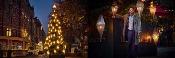 A warmly lit Christmas tree adorned with lantern-style decorations stands elegantly outside The Connaught in London, with a man standing beside it, embracing the festive ambiance.