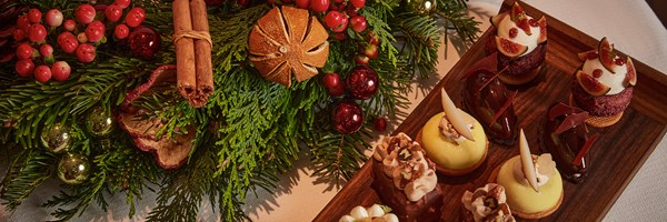 A selection of intricate, festive desserts elegantly arranged next to a beautifully decorated Christmas wreath with berries, pine, cinnamon sticks, and dried citrus slices.