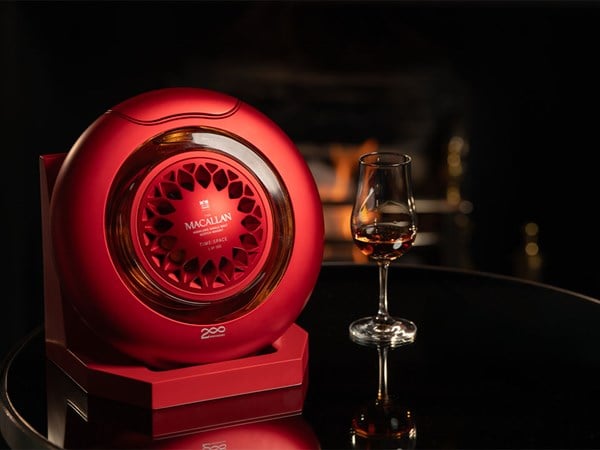 The Macallan Time Series 200 presented in a striking red circular decanter beside a glass of whisky, set against a cozy fireplace backdrop.