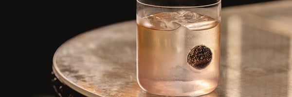 A refined cocktail with a large ice cube and a single dark garnish, served in a minimalist glass on a softly illuminated surface.