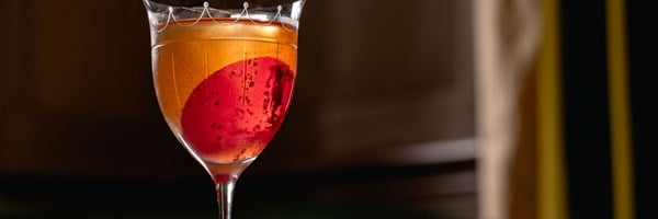 A vibrant amber cocktail with a striking red sphere garnish, elegantly presented in a delicate glass against a dark, sophisticated background.