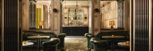 Wide-angle view of The Connaught Bar, showcasing its symmetrical layout, rich textures, dark green banquettes, marble-topped tables, and the opulent mirrored back bar set against ornate panelling.