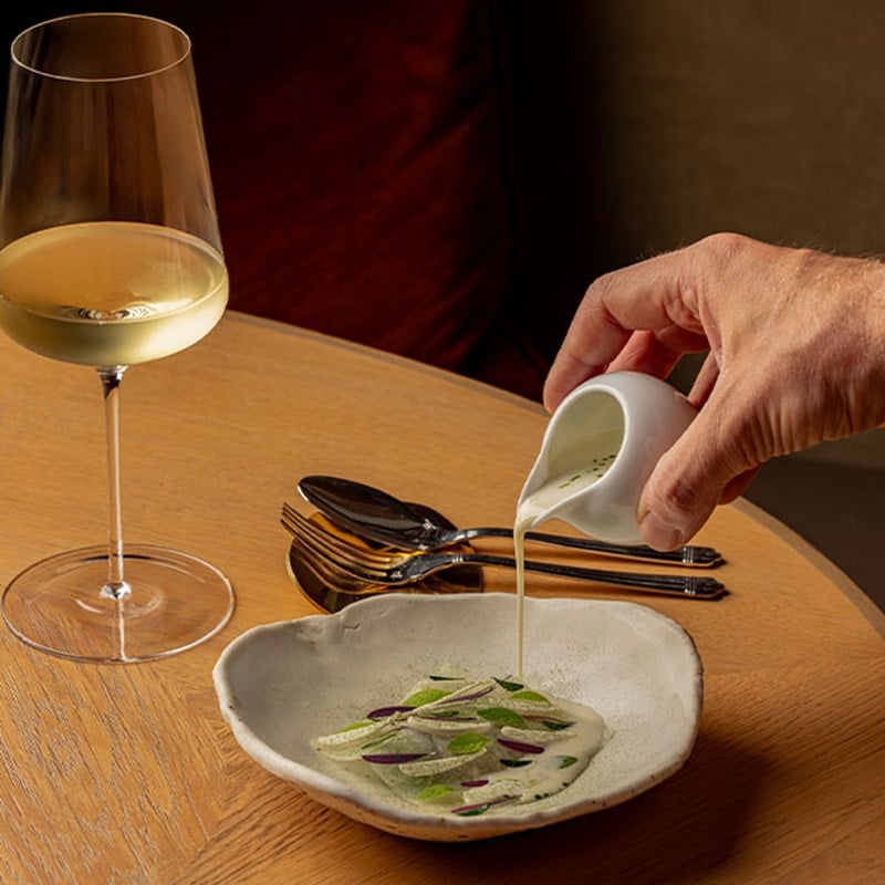 Hand pouring creamy soup into a bowl garnished with thinly sliced vegetables and herbs, with a glass of white wine beside it.
