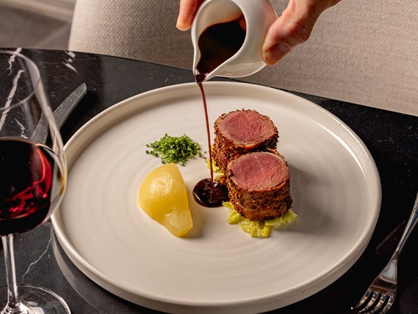 A gourmet plate featuring two medallions of roasted meat crusted with herbs and spices, served with a pear wedge, a dollop of finely chopped herbs, and a pool of rich sauce being poured over the dish, accompanied by a glass of red wine on a black marble table.