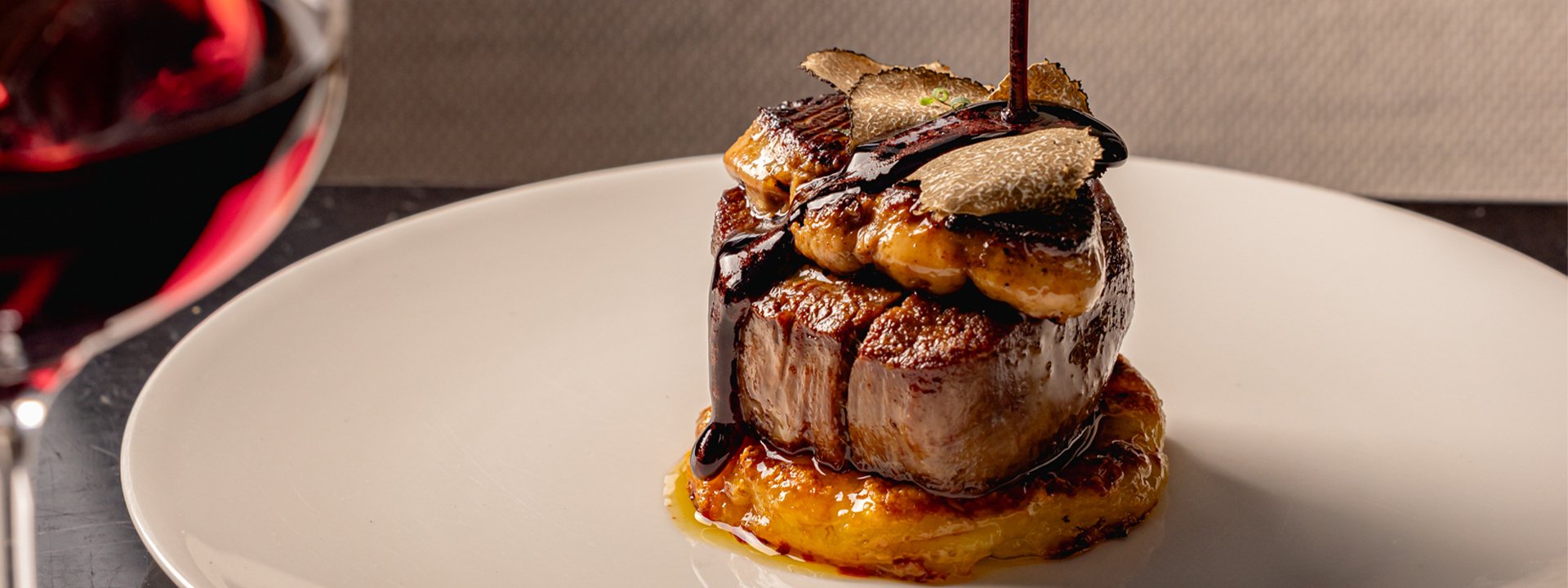 A beautifully plated dish featuring a succulent cut of meat topped with seared foie gras, thin slices of truffle, and a rich glaze, accompanied by a glass of red wine, creating an indulgent and refined dining experience.