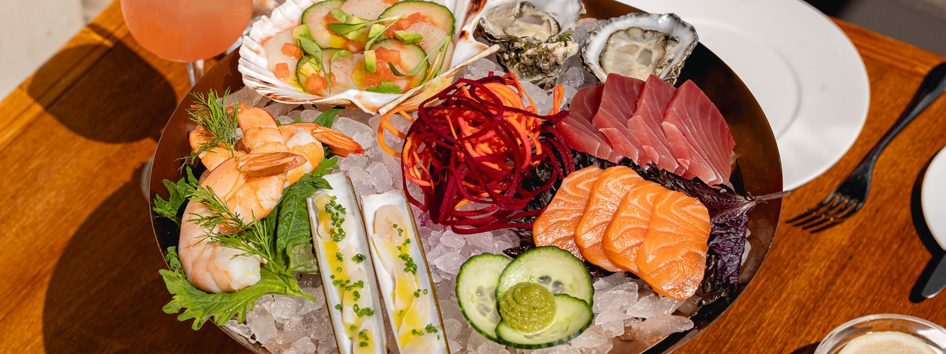 A beautifully arranged seafood platter featuring fresh oysters, shrimp, sashimi, and various shellfish, garnished with vegetables and herbs, served on a bed of ice.