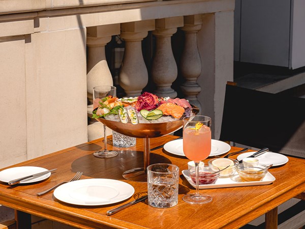 A wooden table is set with two empty plates, a large bowl of seafood on ice, a pink drink in a wine glass, and condiments in small dishes. The scene is outdoors, featuring a stone balustrade in the background.