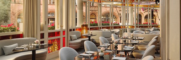 Bright and elegant dining room at Jean-Georges at The Connaught, featuring curved banquettes, soft grey seating, black marble tables, and floor-to-ceiling windows with stained glass details overlooking Mount Street.