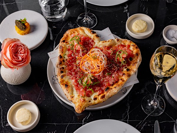 A heart-shaped gourmet pizza topped with finely diced tuna, garnished with delicate ribbons of vegetables, is elegantly presented on a black marble table, accompanied by a glass of champagne, butter, and a refined amuse-bouche.