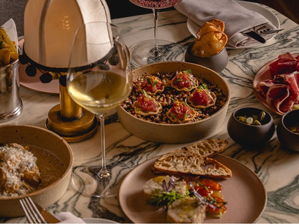 A luxurious spread featuring a glass of white wine, a bowl of tuna tartare bites, charcuterie, fresh bread, and truffle pasta, elegantly arranged on a marble table illuminated by a warm table lamp.