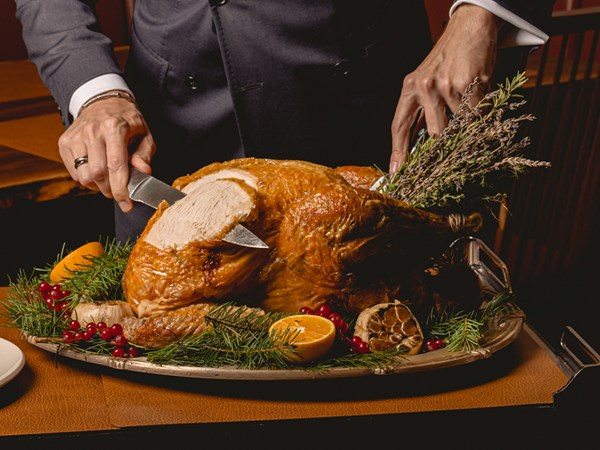 A finely dressed individual carves a golden, herb-roasted turkey garnished with sprigs of rosemary, fresh cranberries, and vibrant orange slices, elegantly presented on a festive platter.