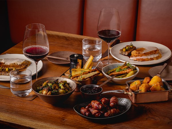 A luxurious festive Sunday lunch spread featuring a variety of dishes, including roasted vegetables, pigs in blankets, crispy potatoes, and an array of meats, accompanied by red wine in an elegant dining setting.