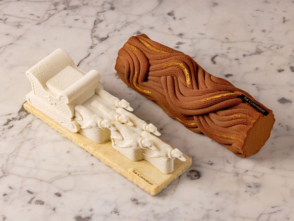 A luxurious Christmas-themed dessert display featuring an intricate sleigh design and a textured Yule log, adorned with gold accents, created by The Connaught Patisserie, resting elegantly on a marble surface.