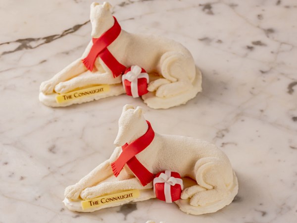 Two elegant white chocolate sculptures of reclining hounds, adorned with vibrant red scarves and accompanied by festive red-and-white gift boxes, crafted by The Connaught Patisserie and displayed on a marble surface.