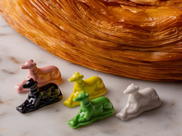 Five colorful porcelain figurines of reclining greyhounds are placed next to the galette, highlighting the pastry's perfectly layered crust.