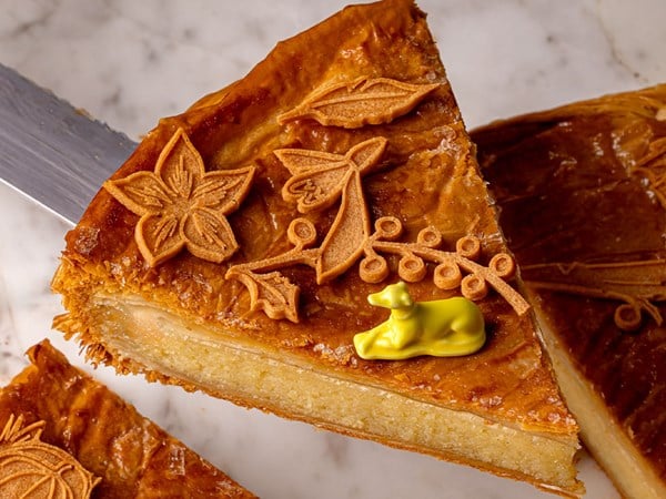 A slice of the galette reveals its almond filling, with a glossy crust and a vibrant yellow greyhound figurine hidden inside.