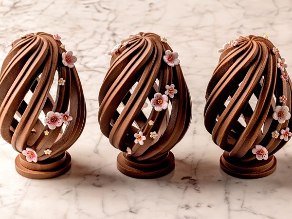 Three intricately sculpted chocolate Easter eggs adorned with delicate pink and white sugar blossoms on a marble surface.