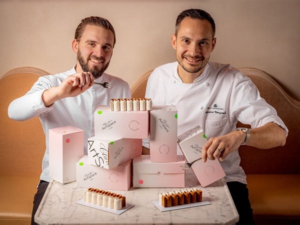 Two chefs proudly present their elegant pastries, surrounded by pastel pink ArtSpace boxes, seated at a marble table.