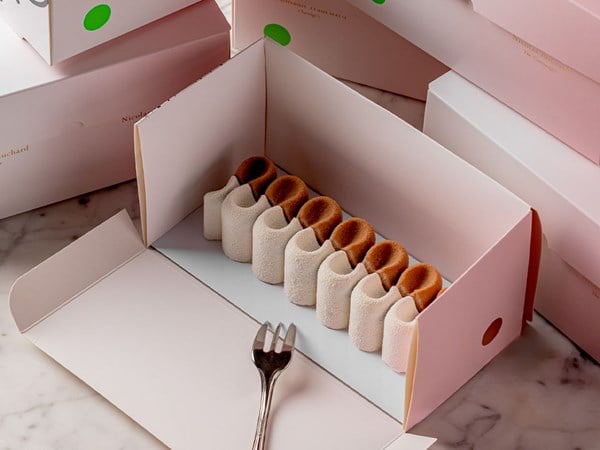 A beautifully designed pastry with a sculptural, wave-like texture sits inside a pastel pink ArtSpace box, accompanied by a silver fork.