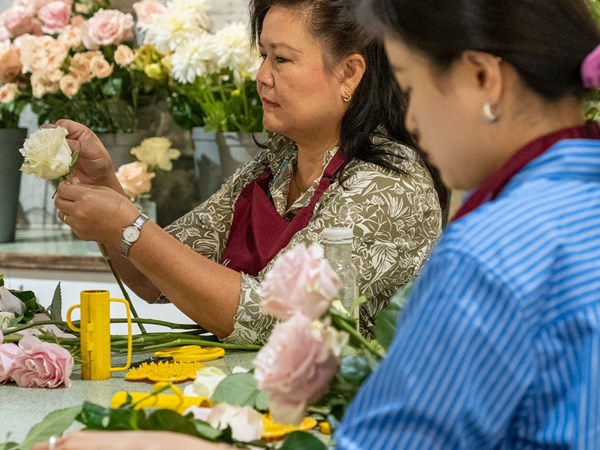 Two women, both wearing maroon aprons, focus intently on crafting floral arrangements; one holds a white rose delicately, while surrounded by an array of pastel-colored flowers in a bustling workshop.