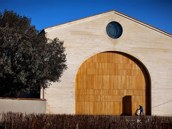 A modern winery facade with a large arched wooden door, light stone walls, and a circular window, with a person in blue rolling a wine barrel under clear blue skies.