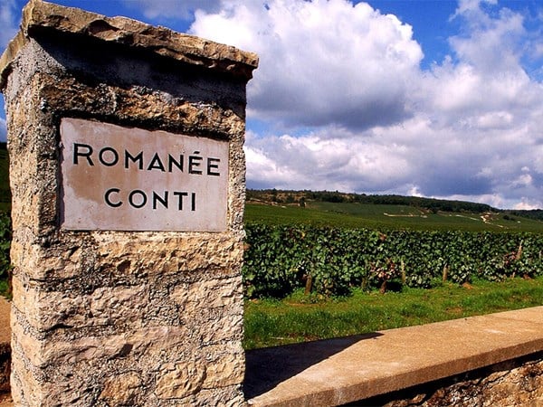 A view of the Romanée-Conti vineyard sign, situated on a stone pillar, with lush green vines stretching across the rolling landscape under a partly cloudy blue sky.