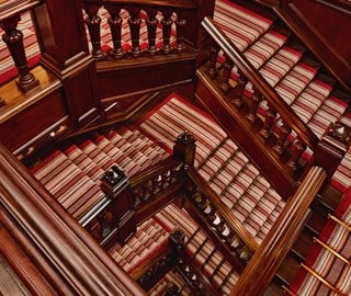 Ornate wooden staircase with striped red and beige carpet runners, viewed from above in a multi-level interior.