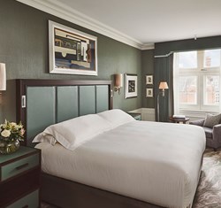 Double bed with white bedding, green eadboard, green bed side tables with a vase of flowers. Beside the bed there is an arm chair and coffee table with books.