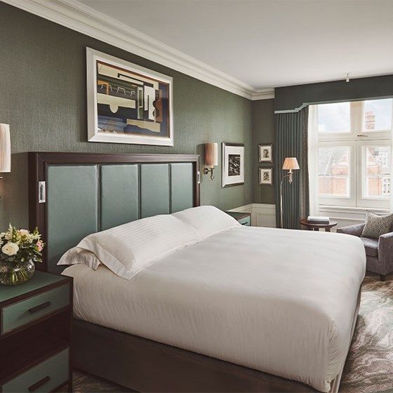 Double bed with white bedding, green eadboard, green bed side tables with a vase of flowers. Beside the bed there is an arm chair and coffee table with books.