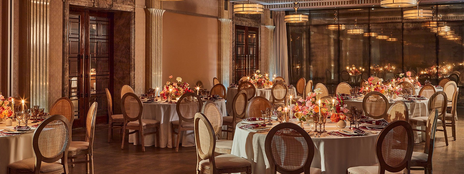 A luxurious dining room is elegantly set for an event, featuring round tables with white tablecloths, floral centerpieces, and candlelight, surrounded by stylish chairs with wicker backs.