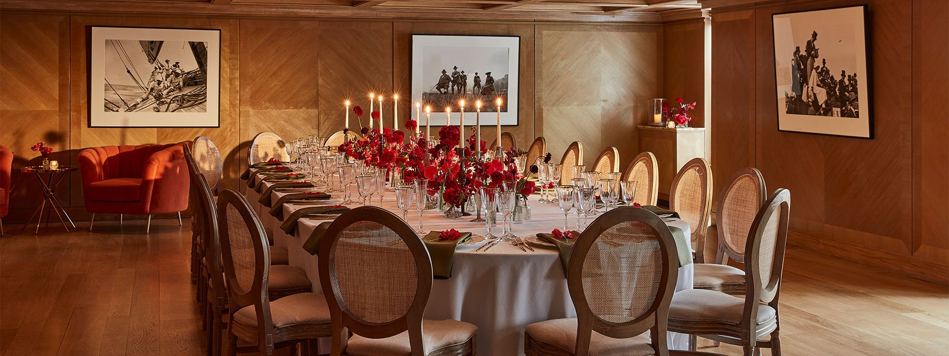 A luxurious dining room is set for an event featuring a long oval table with a white tablecloth, a red floral centrepiece and tall candles. In the background to the left, a round coral armchair is placed next to a round table with a vase and flowers.
