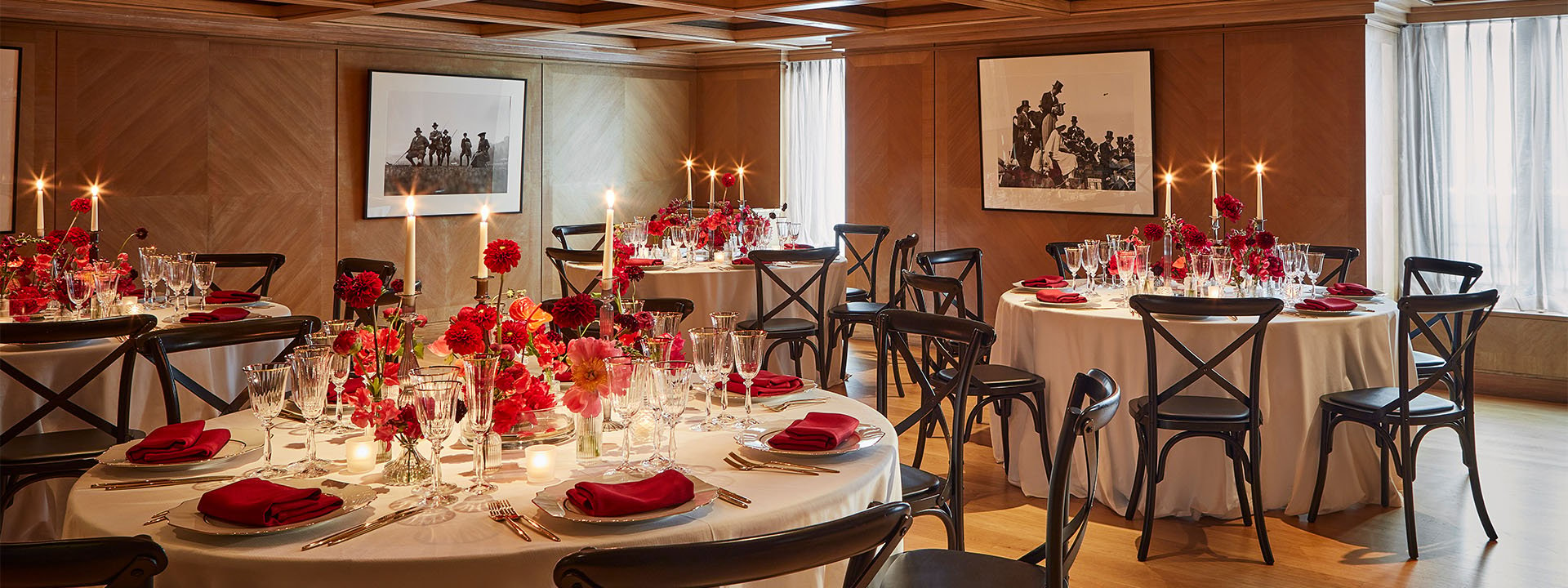 A luxurious dining room is elegantly set for an event, featuring round tables with white tablecloths, red floral centrepieces and candles surrounded by dark wooden chairs. Framed black and white photographs hangs on the wood-paneled walls.