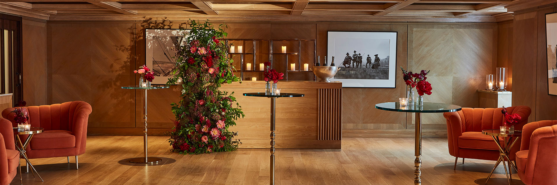 Event room with wooden floor, walls and ceiling, with a couple of small high tables with glass tops/ At the back is a bar with a floral arrangement and several calendars.