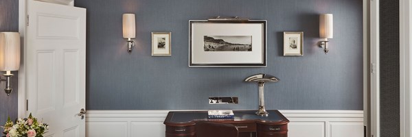 View of a wall with blue wallpaper and black and white images on the wall, framed two lamps attached to the wall. A wooden desk faces the wall, with a brown chair tucked behind it.