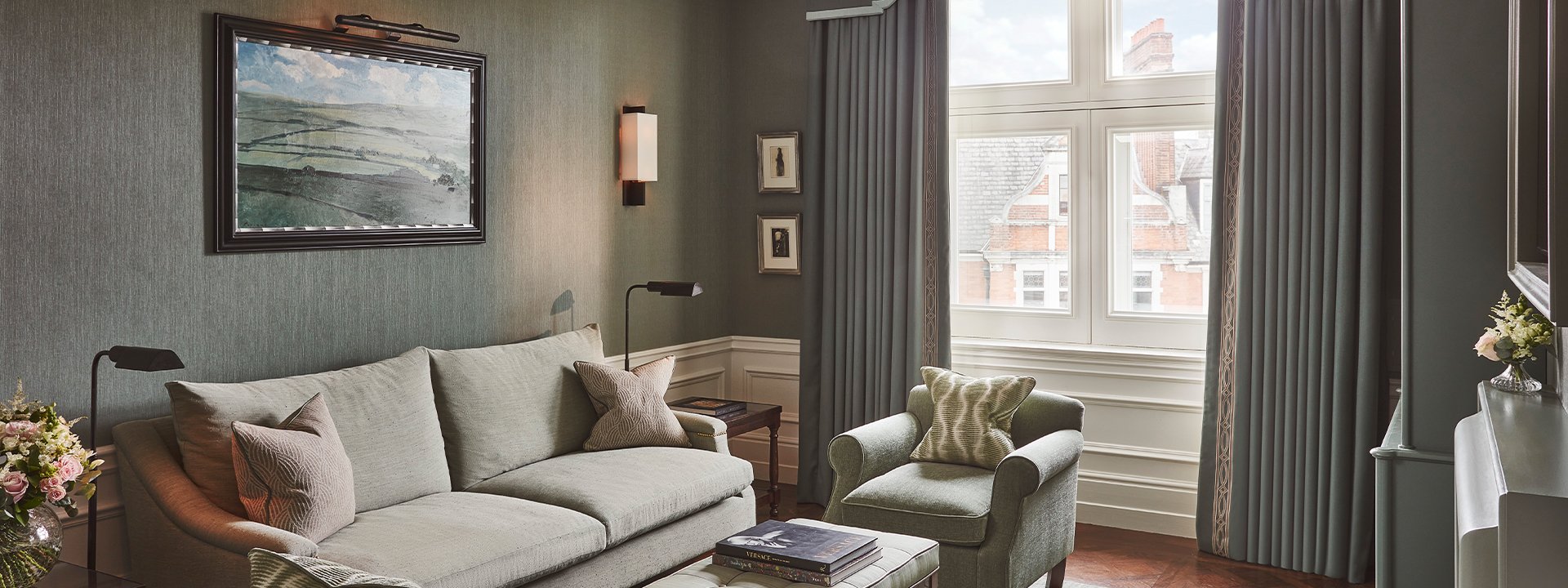 Coburg Suite: Living area of suite with sofa and cushions on the left, and a painting on the wall. A coffee table with books in front of the sofa, and a chair with a green cushion. The colours are shades of green.