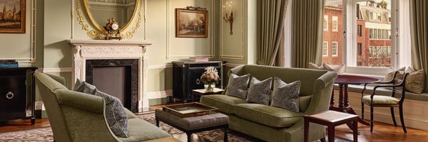 Elegant drawing room with green velvet sofas, a decorative fireplace, and tall windows overlooking red-brick townhouses.