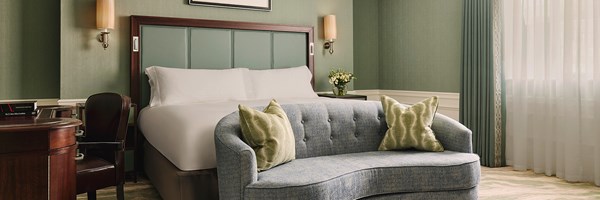 A serene hotel room featuring a plush king-sized bed with a soft blue tufted sofa at its foot, adorned with elegant green cushions and framed by soft, muted green walls.