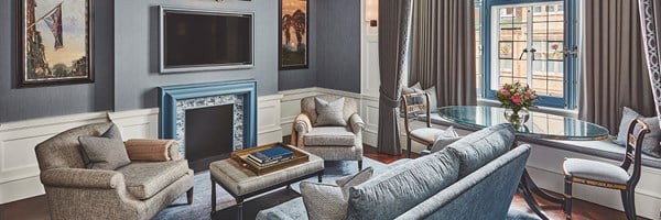 A gracious living space featuring a cozy fireplace with blue and white tiles, luxurious seating, and a charming round table by the window, all set against soothing blue-gray tones for a refined yet inviting ambiance.