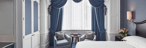 A beautifully appointed bedroom featuring floor-length blue curtains, a stately bed with plush bedding, and two chic armchairs, all arranged to offer a tranquil and elegant setting bathed in natural light.