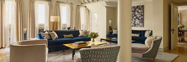 Refined living room with deep blue velvet sofas, contemporary armchairs, and floor-to-ceiling drapes, bathed in natural light beneath a vaulted ceiling.